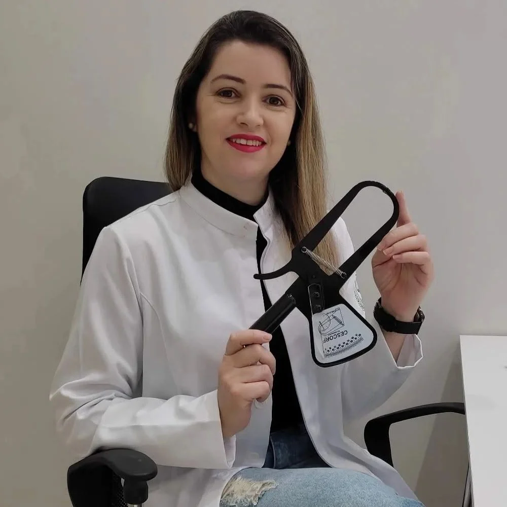 Gisleide de jaleco, sentada no consultório, mostrando o adipômetro usado na avaliação