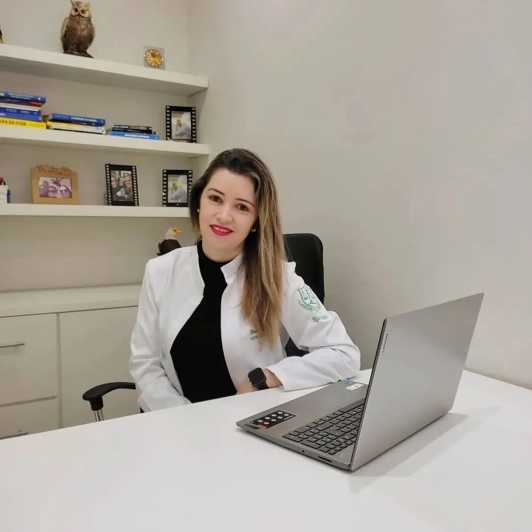 Gisleide sorrindo à mesa do consultório, de jaleco, com o notebook aberto e uma estante de livros ao fundo